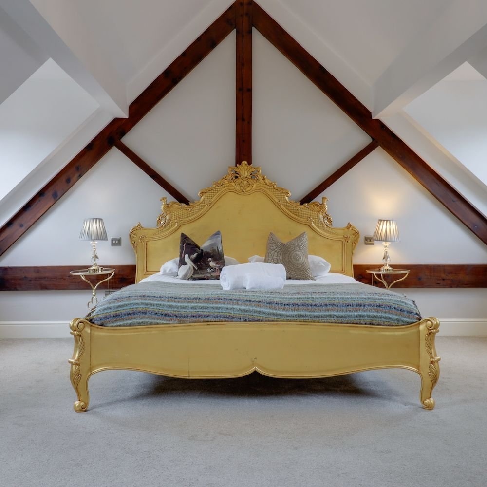 A bedroom set under the eaves with exposed dark timber trusses and an ornate gilt bed frame, flanked by two lamps