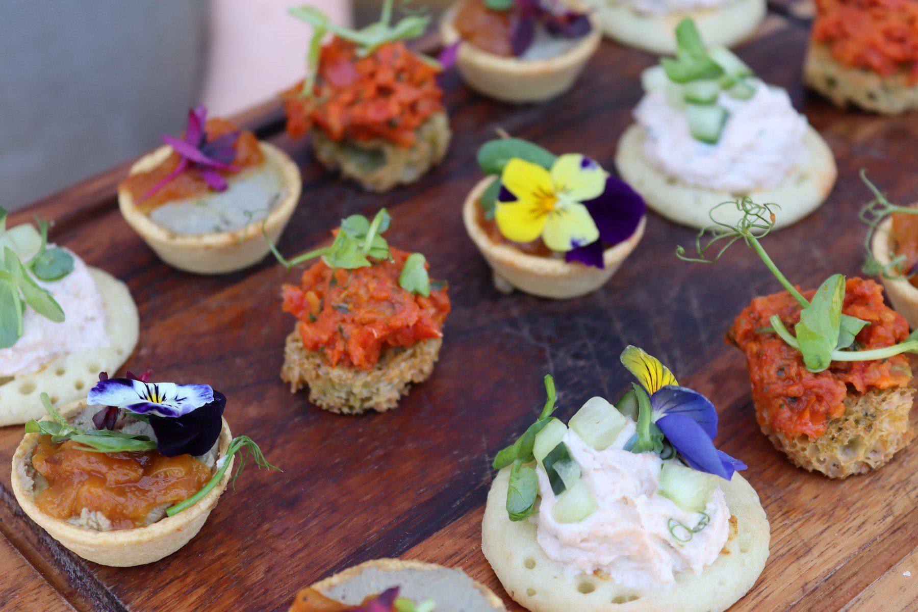Canapés laid out on a dark wooden board — blinis with smoked salmon mousse and diced cucumber, tartlets filled with chutney, and seeded toasts topped with red pepper, each finished with pea shoots and edible violas