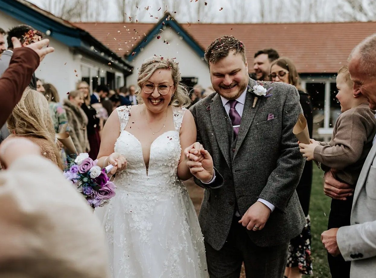 A bride in a lace gown holding a lilac and white bouquet and a groom in a grey tweed suit walking hand in hand through a shower of confetti thrown by their guests outside the Pavilion, both laughing