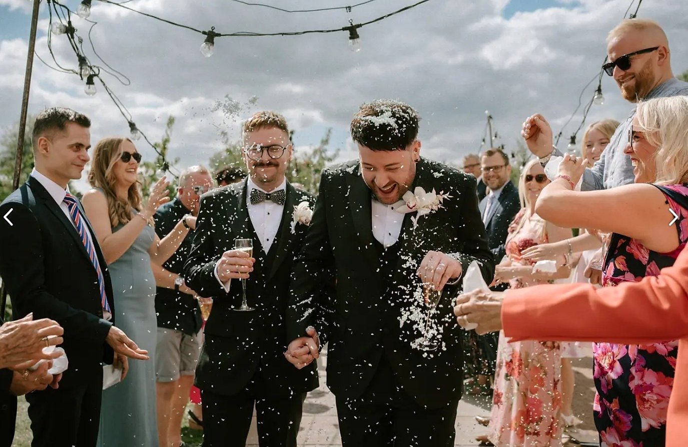 Two grooms in black tuxedos walking hand in hand through a shower of confetti with champagne glasses, their guests throwing petals on both sides beneath festoon lights and a bright sky
