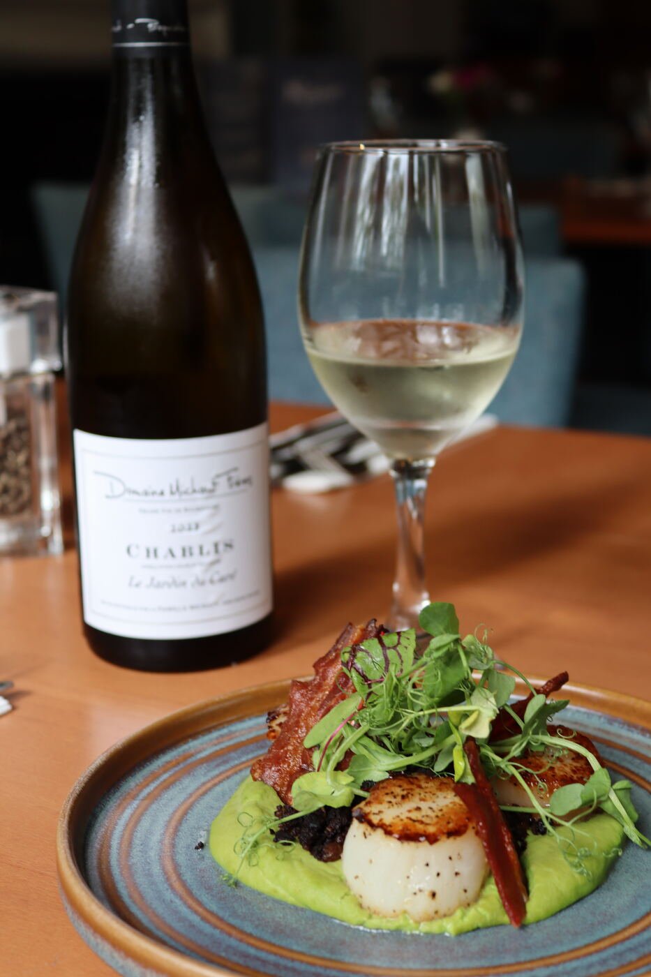 A seared scallop on a bed of green purée with crisp garnish, beside a bottle of Chablis and a glass of white wine