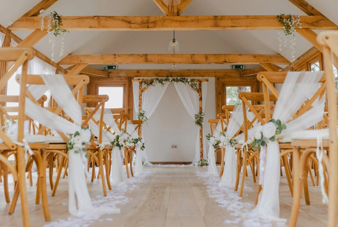 The Glade House set for a ceremony — cross-back chairs dressed in white chiffon and single white roses, an aisle strewn with petals leading to a timber arch hung with white drapes and trailing eucalyptus, wisteria on the oak beams above