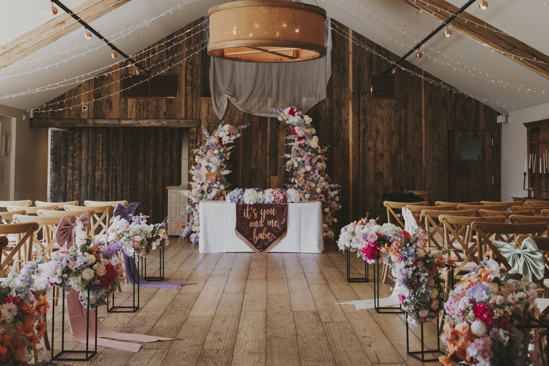 The Glade House set for a ceremony — an aisle of cross-back chairs lined with tall pedestals of pastel flowers, leading to a top table beneath a floral arch and a hanging drape, under fairy lights and a drum shade