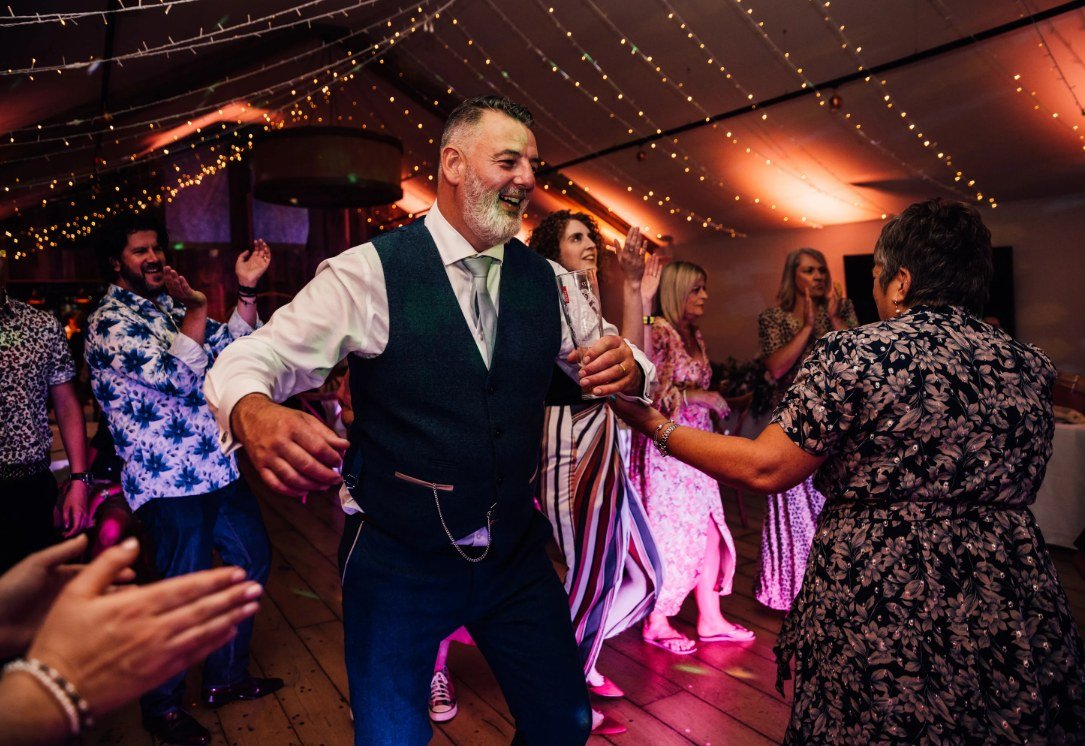 A guest in a waistcoat and sage tie dancing with a drink in hand, laughing, surrounded by other guests mid-dance under fairy lights and warm uplighting in the Pavilion