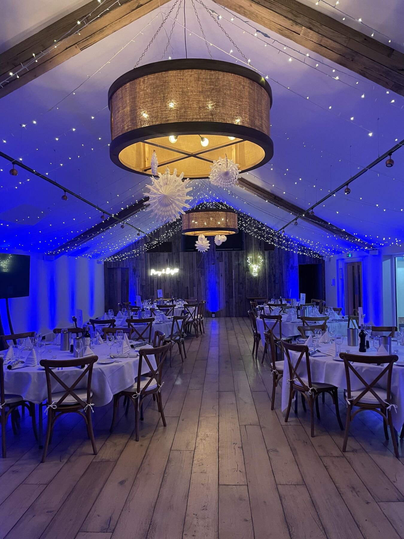 The Pavilion at night lit in deep blue, round tables laid in white linen beneath drum shades and canopies of fairy lights, paper snowflakes hanging from the beams