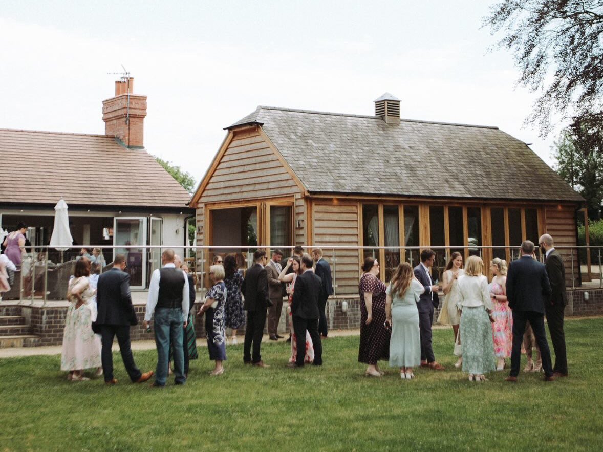 Wedding guests standing in groups on the lawn outside The Pavilion in the sunshine, the oak-clad building and its glazed frontage behind them