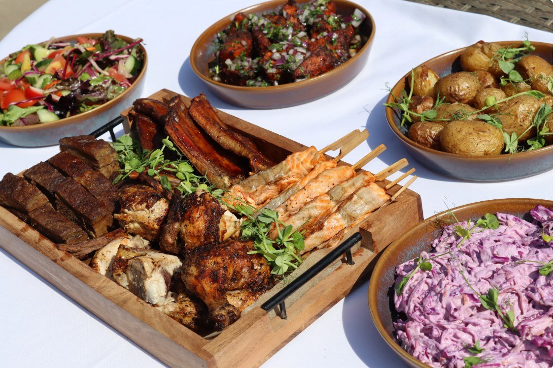 A wooden sharing tray of barbecued short rib, ribs, chicken and salmon and prawn skewers, surrounded by bowls of new potatoes, red cabbage slaw, salad and spiced wings on a white linen table in the sun