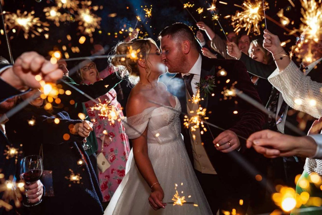 A bride and groom kissing at night inside a tunnel of guests holding lit sparklers, the flares filling the frame around them