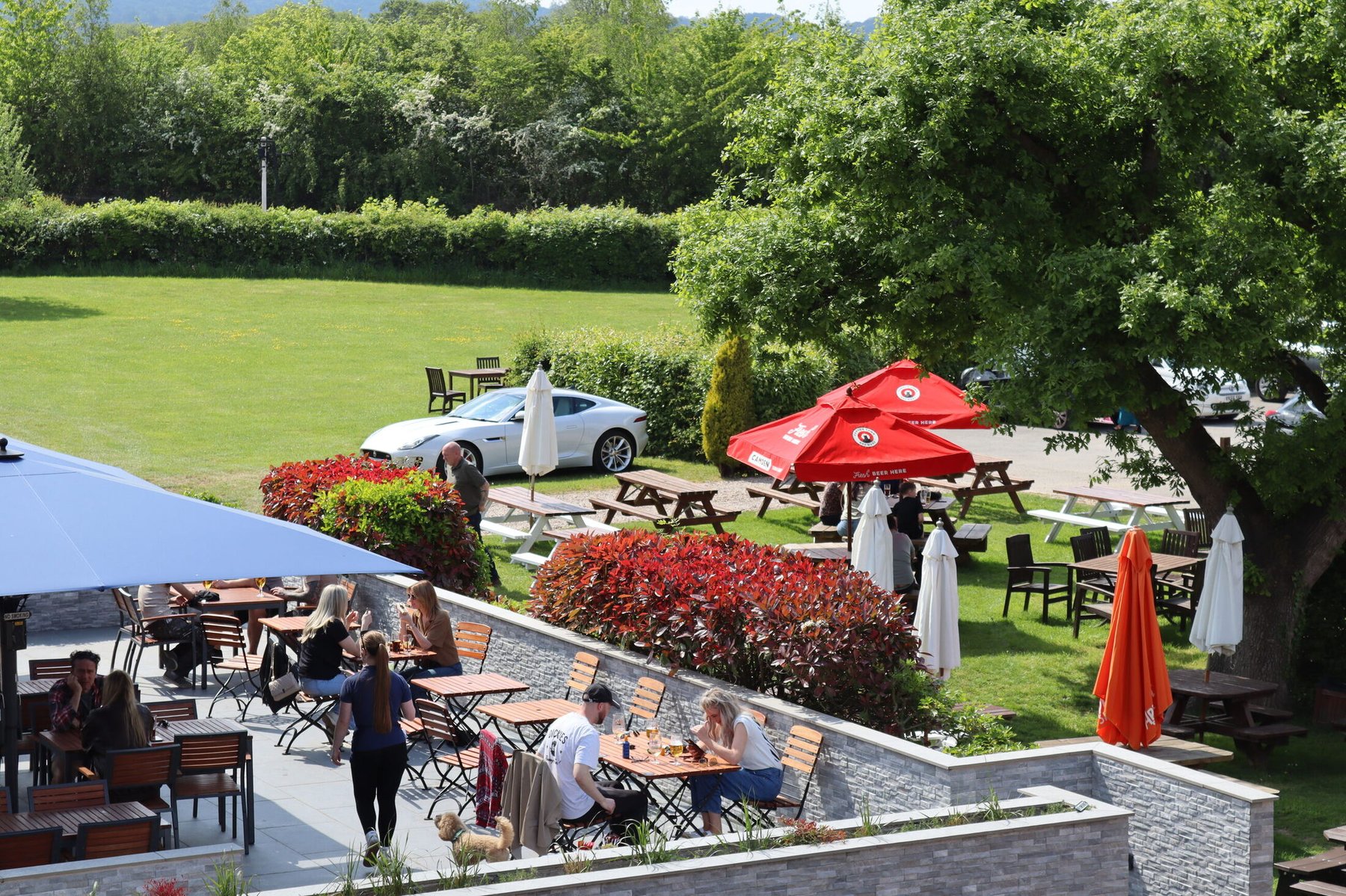 A raised view over the terrace and garden on a sunny day — guests at wooden tables under parasols, picnic benches on the lawn, clipped hedges and open countryside beyond