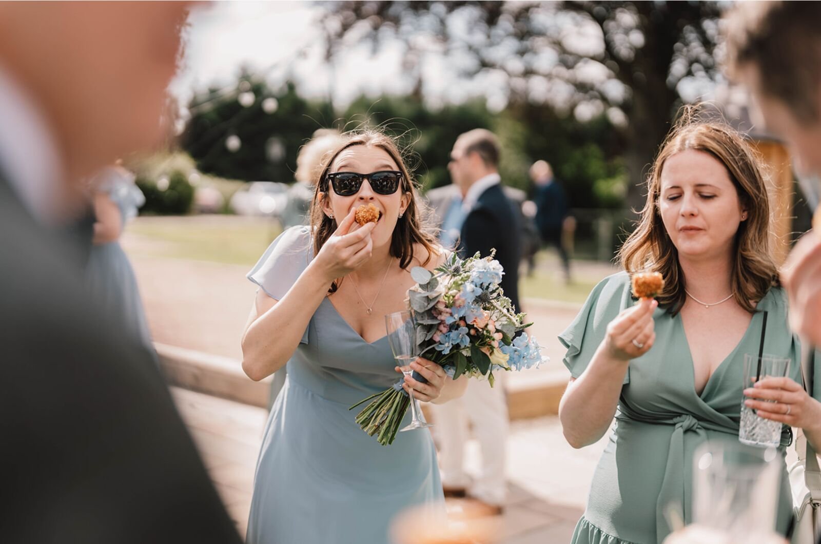 Two guests in sage and blue bridesmaid dresses eating canapés outside in the sunshine, one holding a bouquet and a glass of fizz