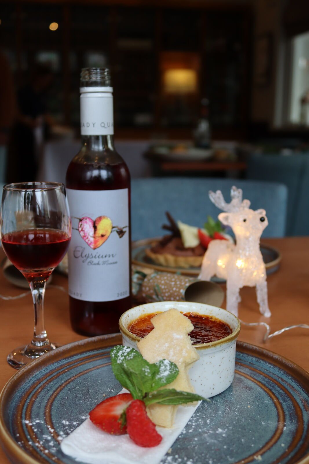 A crème brûlée served with a Christmas tree shortbread and strawberries, beside a bottle and glass of Elysium Black Muscat dessert wine and a light-up reindeer
